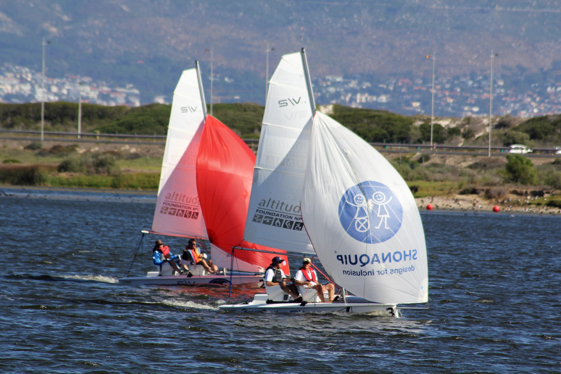 Two SV14 boats sailing on the water with adaptive equipment and sailors with disabilities participating in sailing activities. The boats have distinctive white sails with 'SV' markings and are shown against a scenic backdrop.